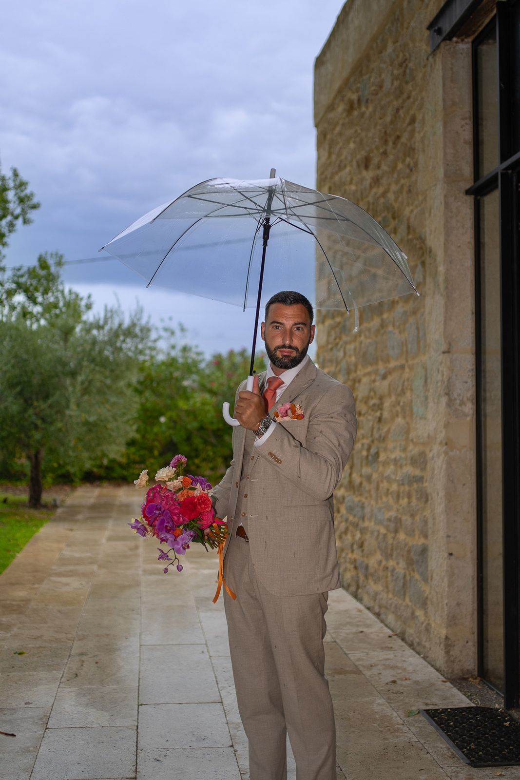 Le marié en costume beige tient le bouquet sous un parapluie transparent, un jour de pluie